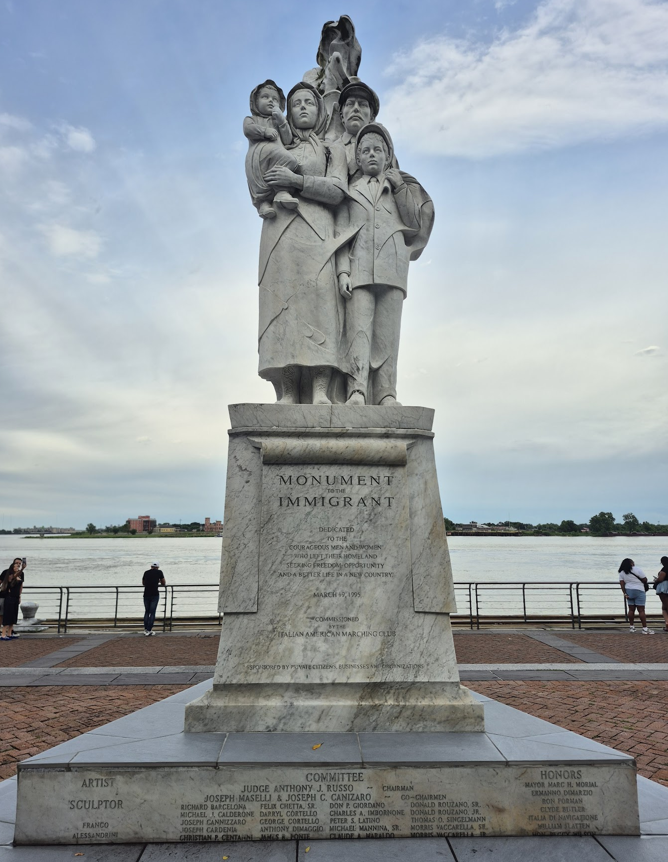 The Monument to the Immigrant in New Orleans — a white marble statue of an immigrant family on a pedestal beside the Mississippi River