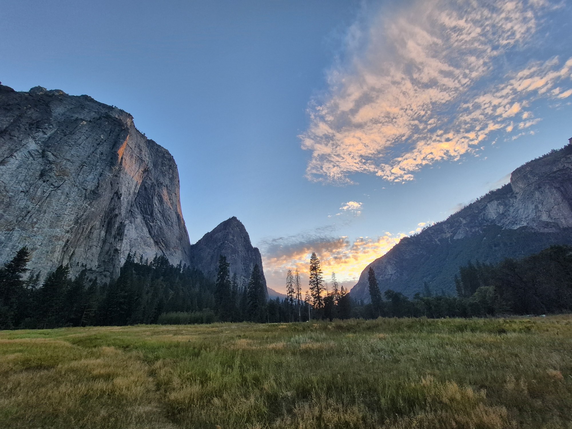Yosemite Valley at sunset from El Capitan Meadow — El Capitan's sheer grey face rising on the left, Cathedral Rocks on the right, a band of orange-lit cloud between them over dry golden meadow grass